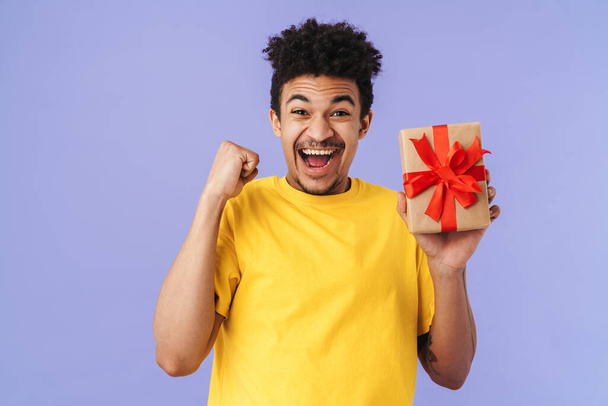 Photo of happy african american man holding gift box and making winner gesture isolated over purple background - Foto, afbeelding