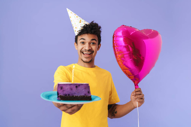 Photo of happy african american man in party cone holding cake and balloon isolated over purple background - Φωτογραφία, εικόνα