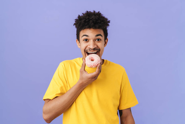 Photo of cheerful african american man eating donut and smiling isolated over purple background - Fotografie, Obrázek