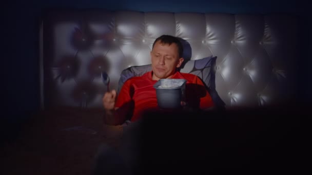 Een jonge man lacht 's nachts op het bed voor de tv met een emmer ijs, de camera beweegt. - Video