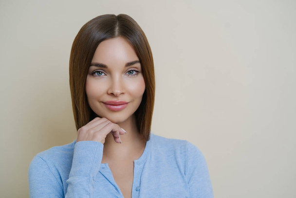 Portrait of beautiful dark haired woman with healthy natural clean skin, touches chin, has manicure and wears blue jumper, isolated over beige background. People, beauty, face expressions concept. - Photo, image