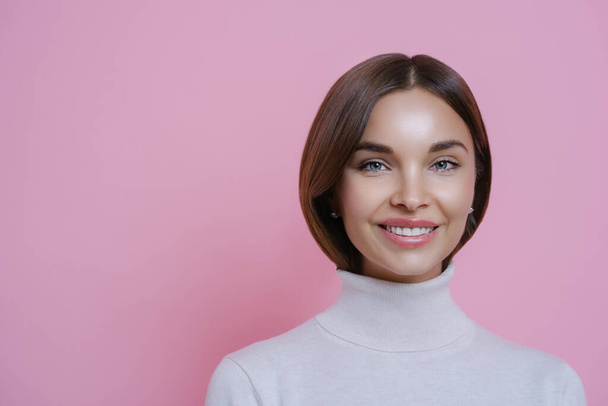 Close up portrait of lovely satisfied joyful woman with dark hair, smiles pleasantly, shows white teeth, wears poloneck, isolated on pink background, empty space for your advertising content. - Фото, изображение