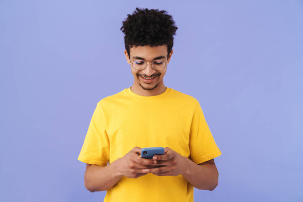 Photo of joyful african american man in eyeglasses smiling and using smartphone isolated over purple background - Photo, Image