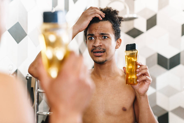 Photo of scared half-naked african american man holding shampoo and looking at mirror in bathroom - Fotografie, Obrázek