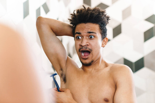 Photo of shocked half-naked african american man shaving his armpits and looking at mirror in bathroom - Foto, Bild