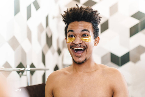 Photo of laughing half-naked african american man with eye patches looking at mirror in bathroom - Fotó, kép