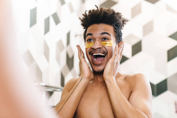 Photo of excited half-naked african american man with eye patches looking at mirror in bathroom - Φωτογραφία, εικόνα