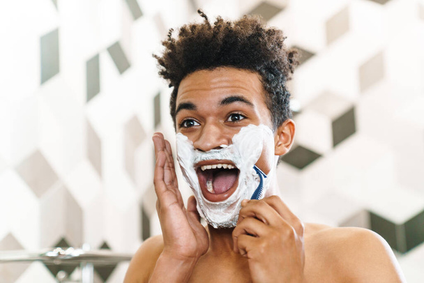Photo of excited half-naked african american man shaving with foam while looking at mirror in bathroom - Фото, изображение