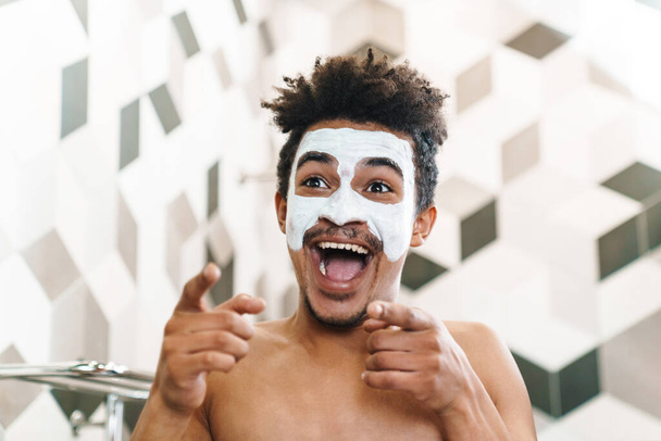 Photo of excited half-naked african american man with cosmetic mask on his face pointing fingers at mirror in bathroom - Фото, изображение