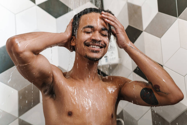 Photo of cheerful half-naked african american man smiling while taking shower in bathroom - Φωτογραφία, εικόνα
