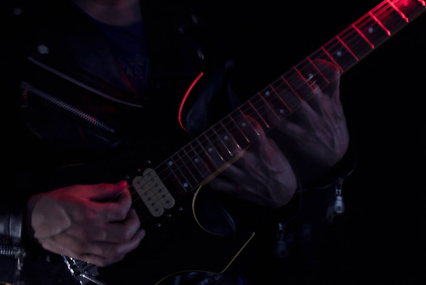  Długa ekspozycja zdjęcie człowieka w skórzanej kurtce grającego na czarno-żółtej gitarze elektrycznej w pomieszczeniach oświetlonych czerwonymi i białymi światłami. Koncepcja rocka i muzyki - Zdjęcie, obraz