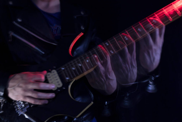 Długa ekspozycja zdjęcie człowieka w skórzanej kurtce grającego na czarno-żółtej gitarze elektrycznej w pomieszczeniach oświetlonych czerwonymi i białymi światłami. Koncepcja rocka i muzyki - Zdjęcie, obraz