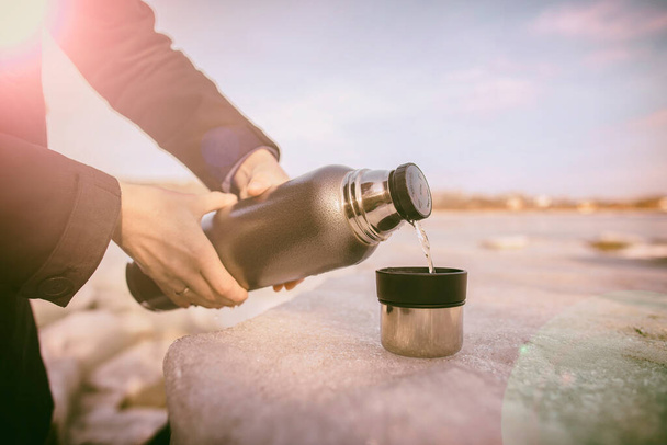 De mens schenkt hete thee uit een thermoskan in een beker die op een ijsschots op de oever van de rivier staat in het voorjaar zonnige dag - Foto, afbeelding