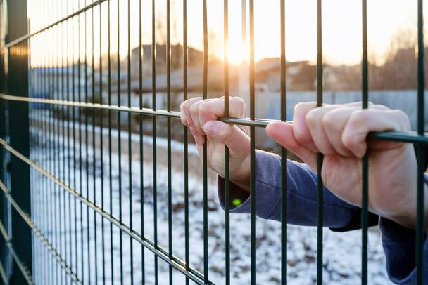 De handen houden het metalen hek vast in de stralen van de ondergaande zon in de winter - Foto, afbeelding