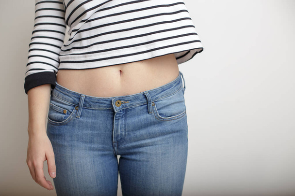 Young girl in blue jeans and a striped sweater and raised her hand bare belly with a beautiful waist and umbilicus - Фото, зображення