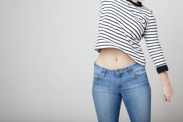 Young girl in blue jeans and a striped sweater and raised her hand bare belly with a beautiful waist and umbilicus - Foto, Imagen