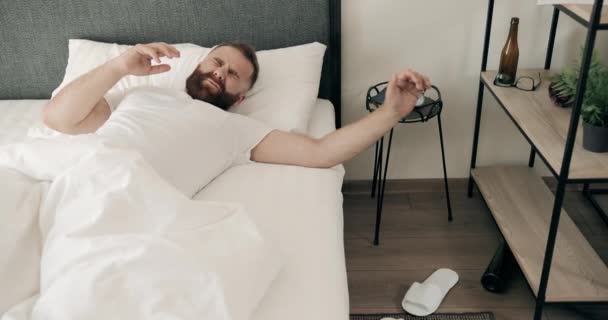 Tired bearded young man waking up in bed and having hangover with head pain. Handsome adult guy in 30s grimacing and touching head with both hands. Concept of bad morning. - Кадры, видео