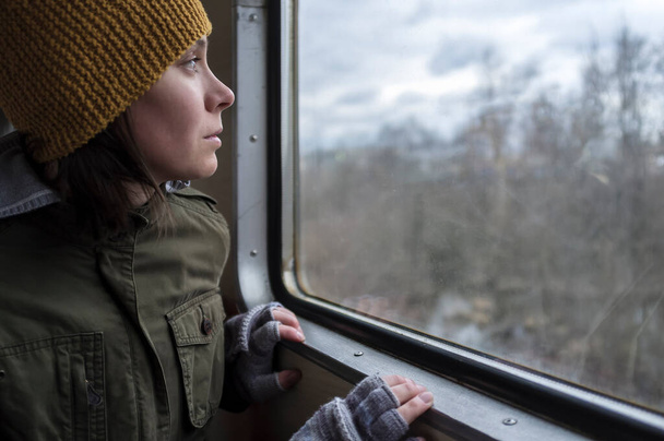 Schöne Mädchen mit einem Wollmütze reist mit dem Zug und schaut aus dem Fenster in den Himmel in den Wolken und Frühlingswald. - Foto, Bild
