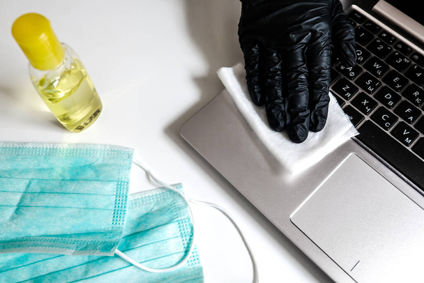 Limpar laptop com toalhetes úmidos antibacterianos para evitar a propagação do vírus e matar bactérias. Medidas de segurança pessoal em casa durante o isolamento vívido. Garrafa desinfetante, máscaras na mesa branca perto do computador - Foto, Imagem