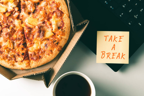Pizza in box, cup of coffee, laptop and sticker with text TAKE A BREAK on table in office. - Foto, imagen