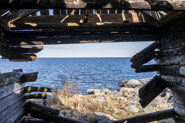 Oude houten hut, door de verwoeste muur is zichtbaar de rotsachtige kust met vegetatie en het meer. Lente, een zonnig landschap. - Foto, afbeelding