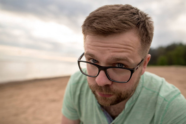 Homme barbu à lunettes regarde les yeux bleus douteux fronçant les sourcils, sur la plage de sable fin de la mer par une soirée nuageuse. - Photo, image