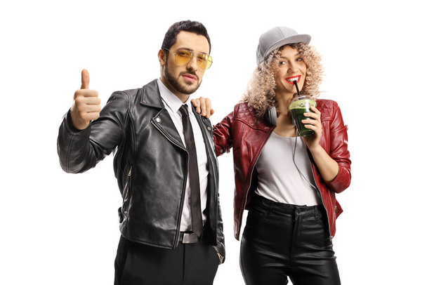 Trendy man with sunglasses showing thumbs up and a woman in leather jackets drinking a green smoothie isolated on white background - Photo, image
