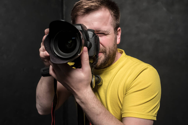 Красивый мужчина - фотограф в жёлтой футболке снимает в студии на DSLR-камеру, которая стоит на штативе. Крупный план
. - Фото, изображение
