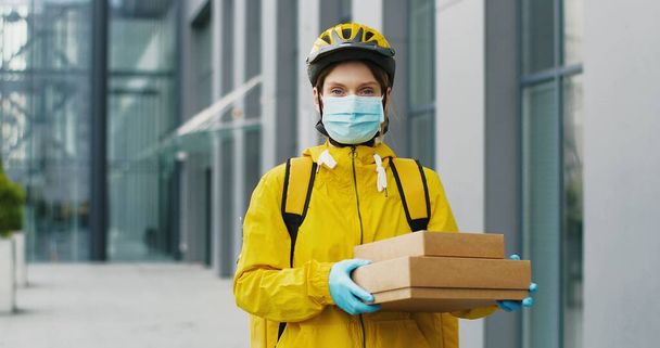 Portrait of young woman delivery worker in medical mask standing at street and holding carton boxes. Pretty female courier with parcels - Valokuva, kuva