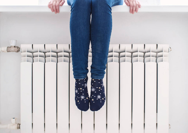 The girl sits on the windowsill and warms her feet in pretty socks at the radiator, on a cold winter day. - Фото, изображение