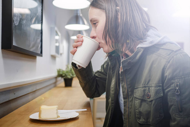 Thoughtful woman drinking cappuccino in a paper cup and eating a cheesecake in a cozy cafe. Close-up. - Photo, Image