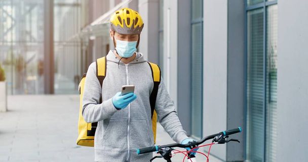 Deliveryman in medical mask, gloves and casque leading his bicycle at street, tapping on cellphone, courier having phone call. - Photo, image