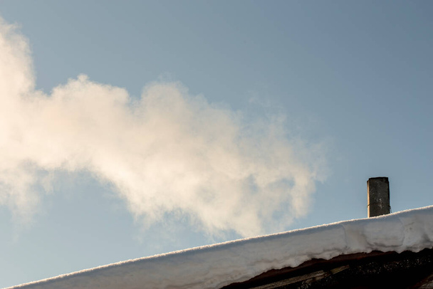 Білий дим зі старого димоходу на даху вкритий снігом, на тлі блакитного неба, раннього морозного, сонячного ранку
. - Фото, зображення