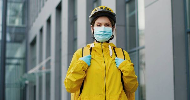 Portrait of beautiful woman delivery worker in medical mask and helmet for riding bike standing at street. Pretty female courier with backpack. Food delivery concept - Foto, Imagen