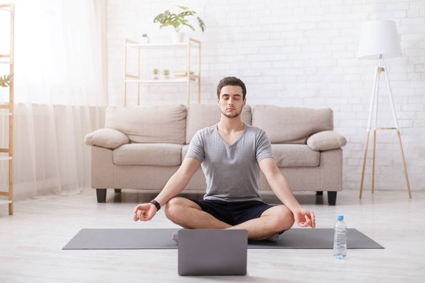 Медитация и йога. Парень с закрытыми глазами сидит в позе лотоса на коврике, с ноутбуком
 - Фото, изображение
