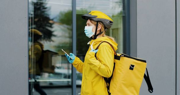 Close up of young woman delivery worker in medical mask standing at street and using smartphone - Zdjęcie, obraz