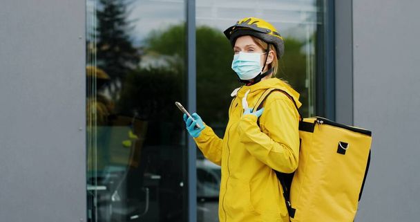 Portret kuriera w masce medycznej i kasku do jazdy na rowerze z żółtą torbą termiczną do dostarczania żywności za pomocą smartfona. Koncepcja dostaw żywności - Zdjęcie, obraz