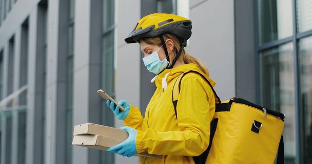 Close up of young woman delivery worker in medical mask standing at street and holding carton boxes and using smartphone - Foto, Imagem
