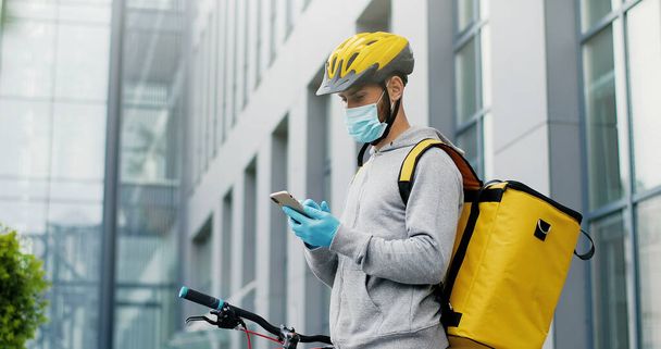 Portrait of male courier in medical mask and helmet for riding bike on bicycle delivering food. Deliveryman using smartphone - 写真・画像