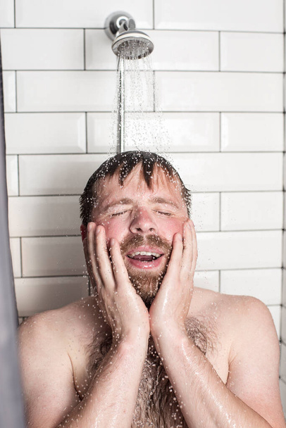 Egy jóképű, szakállas férfi áll a zuhany alatt, meleg víz alatt, fogja a kezét az arcán és boldogan mosolyog.. - Fotó, kép