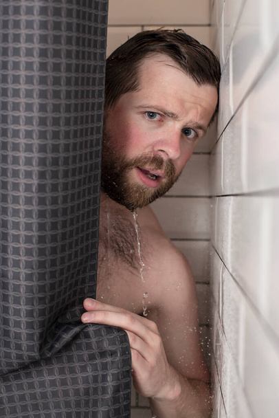 Красивый бородатый мужчина с мокрыми волосами смотрит из-за занавески в душе. Крупный план
. - Фото, изображение