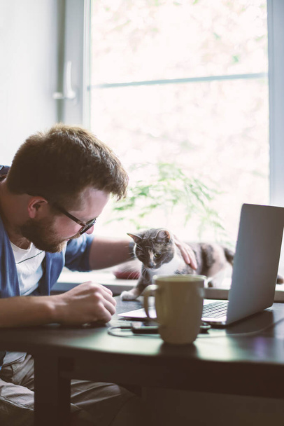 Ein bärtiger Mann mit Brille machte am frühen Morgen eine Pause von der Arbeit mit dem Laptop, um seine geliebte Katze vor dem Fenster zu streicheln.. - Foto, Bild