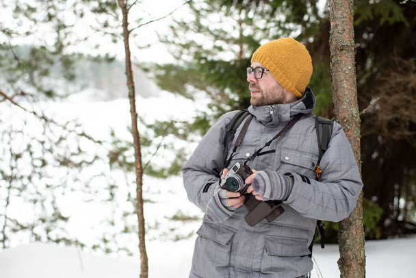 A bearded man with glasses holds a camera in his hands and stands leaning on a trunk of a young pine tree in a winter frosty forest, against the background of a lake, trees and snow. - Photo, image
