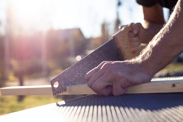 A man cuts a wooden product with a carpentry hand saw, in the sunshine on a warm summer day, outdoors. - Photo, image
