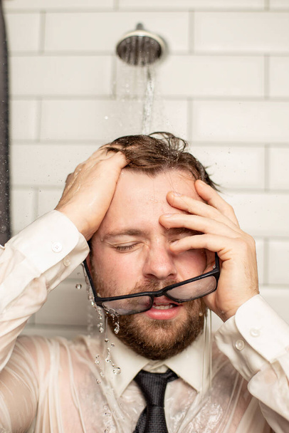 Uomo barbuto sconvolto con occhiali cadenti in camicia da lavoro e cravatta tenendo le mani sopra la testa in piedi sotto la doccia sotto l'acqua corrente calda dopo una dura giornata di lavoro. Primo piano
. - Foto, immagini