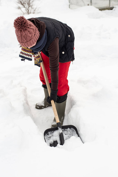 Žena v teplém oblečení pomocí černé plastové lopaty s dřevěnou rukojetí dělá cestu ve sněhu. Detailní záběr. - Fotografie, Obrázek