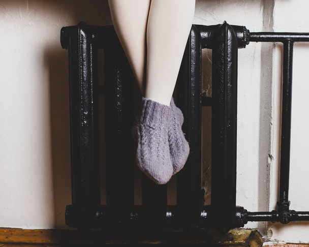 A menina está sentada no parapeito da janela e está aquecendo as pernas em meias de malha de lã no radiador, em uma noite fria de inverno.
. - Foto, Imagem