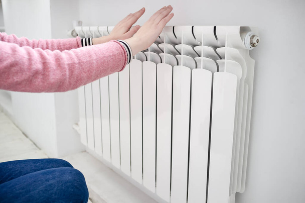 menina em um suéter rosa e jeans azul congelou e aquece as mãos perto do radiador multicell branco
 - Foto, Imagem