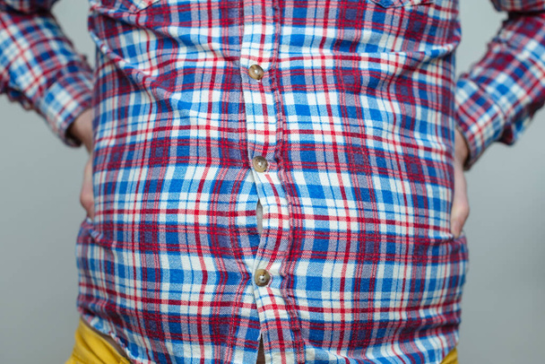 lihava mies ruudullinen paita on vaikeuksia kiinnittää hänen pieniä vaatteita napit; hänen paljain vatsa näkyy reikien läpi - Valokuva, kuva