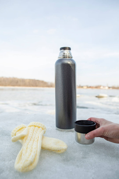 Bir fincan sıcak çay tutan bir kadının eli, üzerinde termos olan bir buz kütlesi kullanarak ve arka planda yün eldivenler uzanarak bahar güneşinde nehri görebilirsiniz.. - Fotoğraf, Görsel